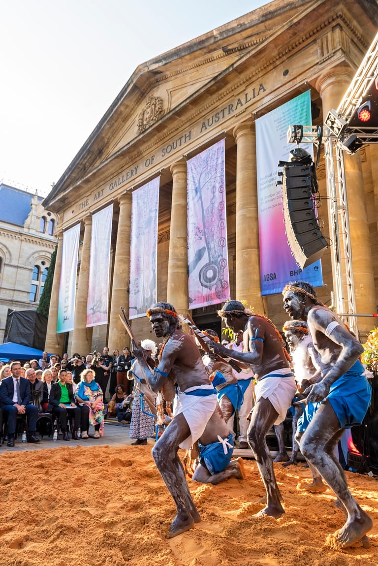Indigenous dancers perform on red earth outside a grand heritage building with classical columns and colourful banners.