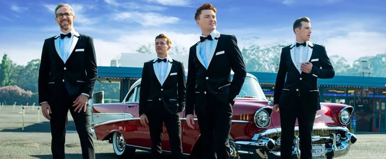 Four men in matching black tuxedos stand in front of vintage red cars and a mountain landscape.