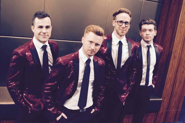 Four men in formal attire, two wearing burgundy sequined jackets, posed against a dark panelled backdrop.