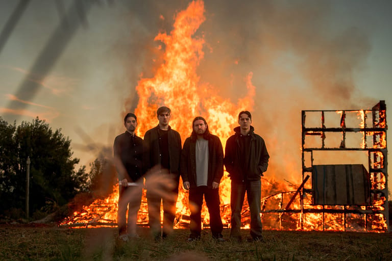 Four figures stand silhouetted against a towering blaze and wooden structures at dusk.