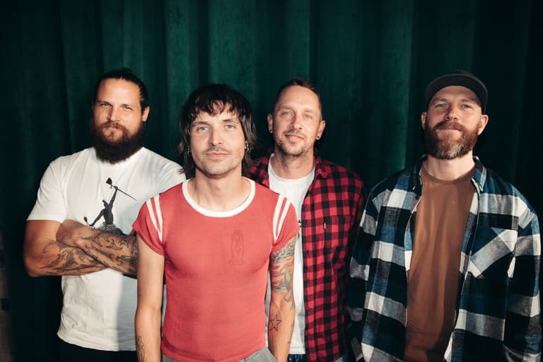 Four bearded men in casual shirts stand together against a dark teal curtain backdrop.