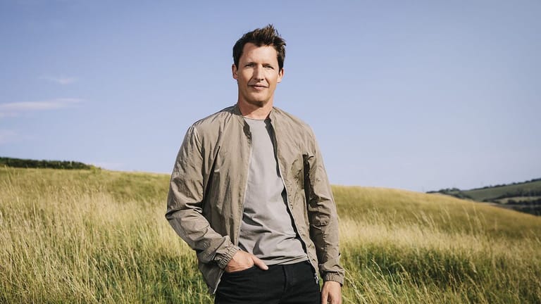 Man in a tan jacket standing in an open grassland field under clear sky.