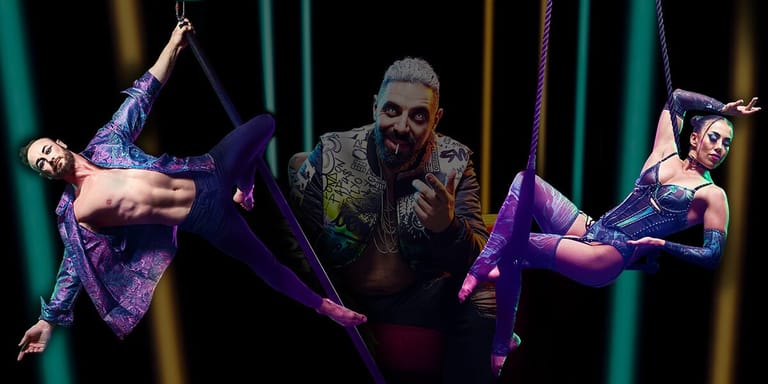 Bearded performer in ornate jacket suspended between two aerial acrobats in purple lighting during a stage production.