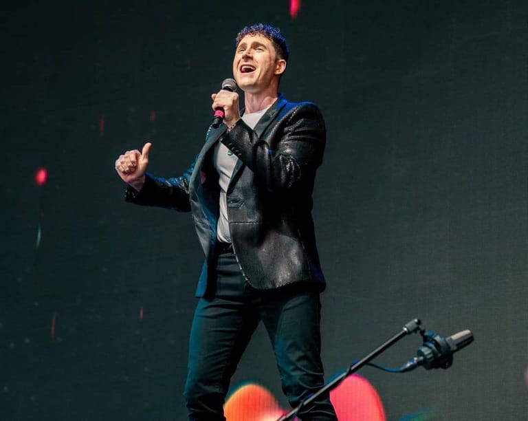 Performer in black leather jacket singing into microphone on stage with coloured stage lights behind.