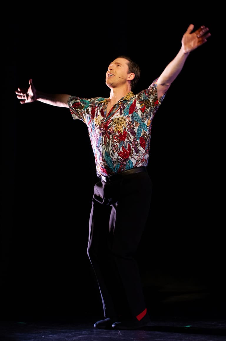Performer in a sequined, multicoloured jacket with arms raised triumphantly on a darkened stage.