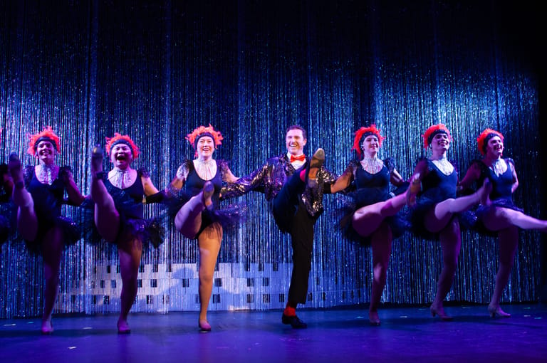 Seven dancers in purple costumes perform a synchronised high-kick routine on a blue-lit stage with vertical backdrop.