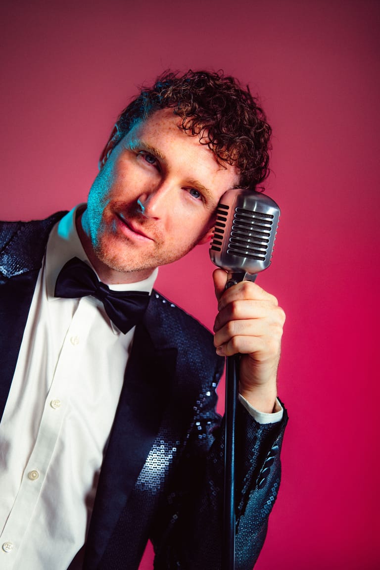 Man in sequined jacket and bow tie holding vintage microphone against pink backdrop.