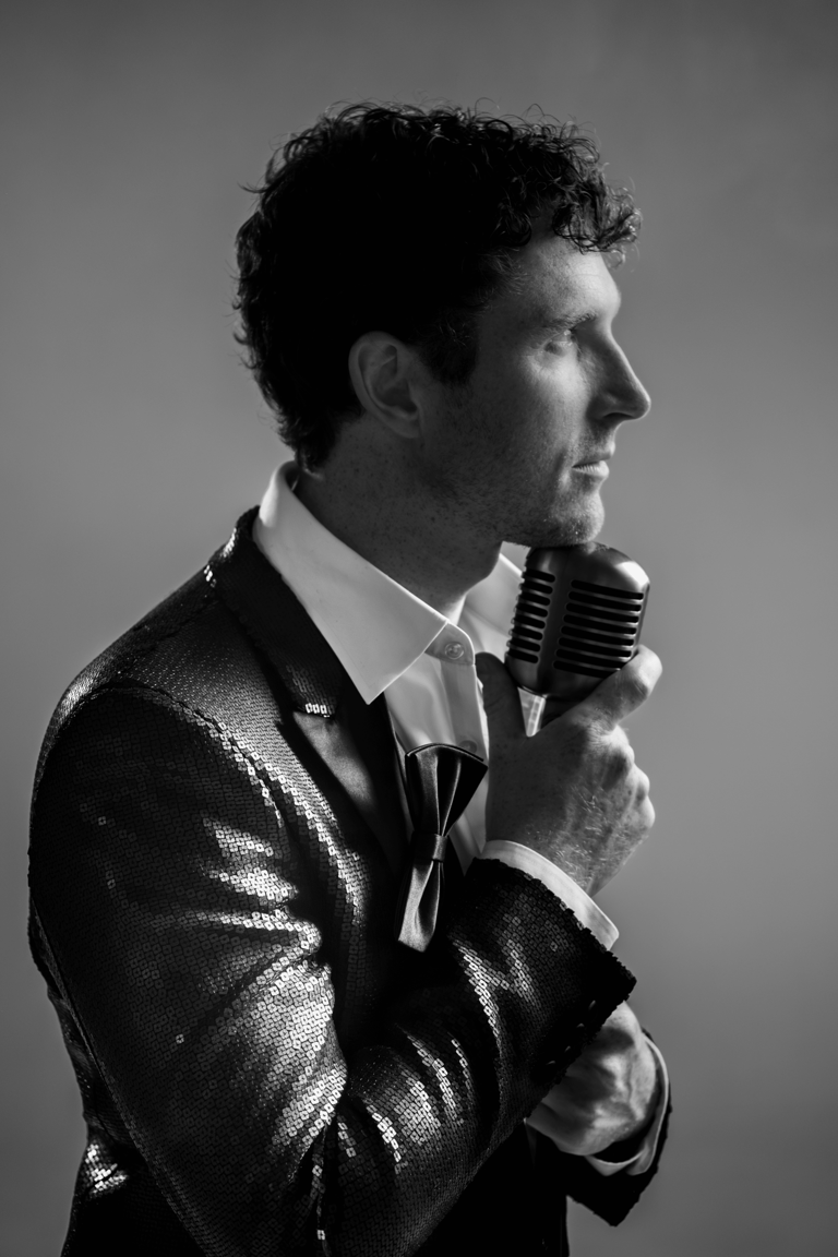 Man in shimmer jacket holds vintage microphone in black-and-white portrait profile shot.
