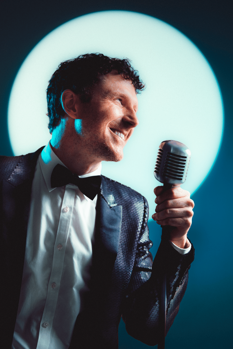 Man in formal suit and bow tie holding vintage microphone against glowing turquoise circular backdrop.