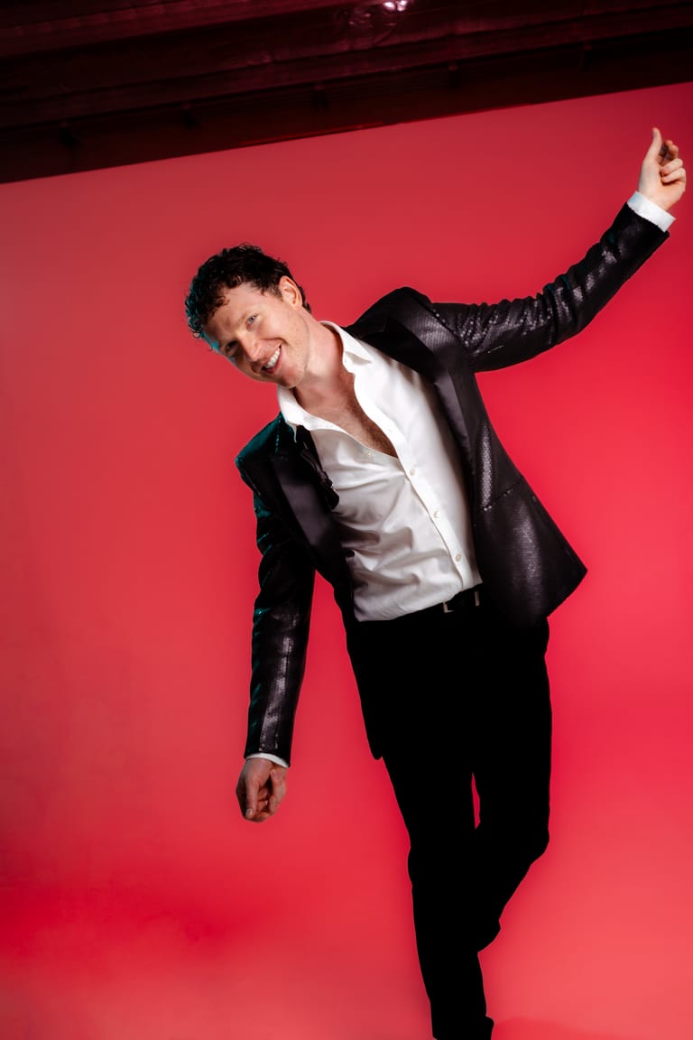 Person in black leather jacket and white shirt striking a dynamic pose against a bright red backdrop.