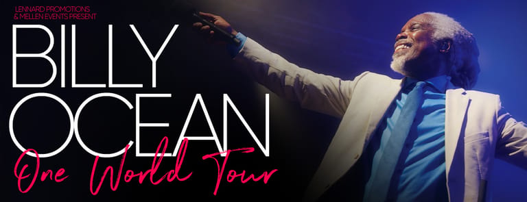 Billy Ocean performs on stage with arm raised, wearing a light-coloured jacket under blue stage lighting.