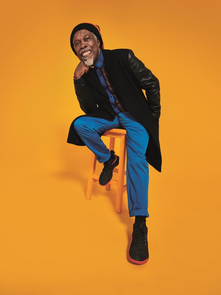 Man in black jacket and blue jeans sitting on orange stool against yellow background, smiling at camera.
