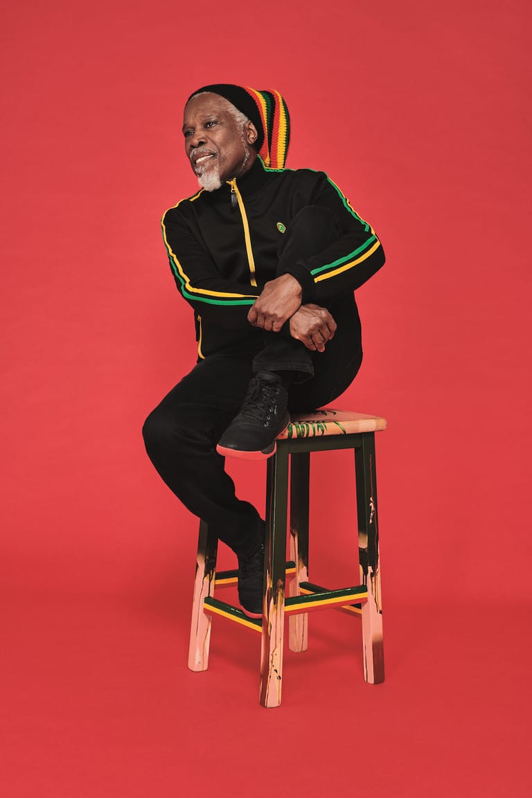 Person in black tracksuit with yellow and green stripes seated on wooden stool against bright red background.