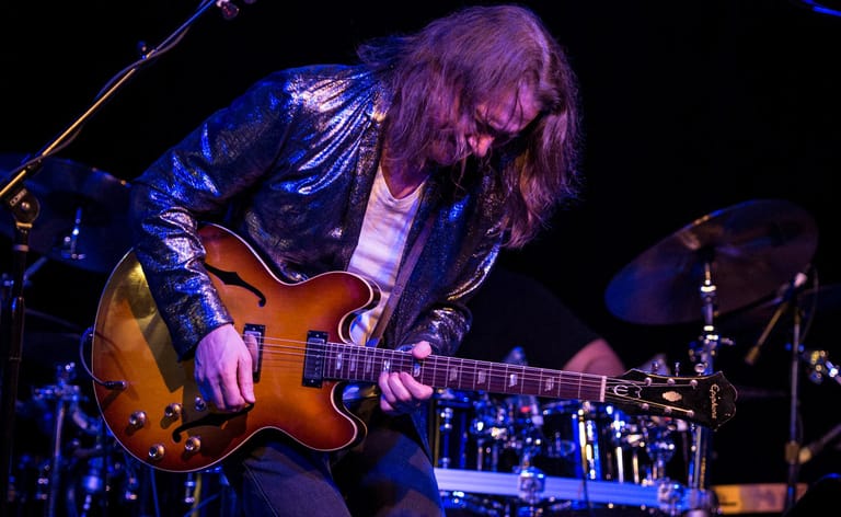 Guitarist with long hair playing sunburst electric guitar on stage, lit by blue spotlights.
