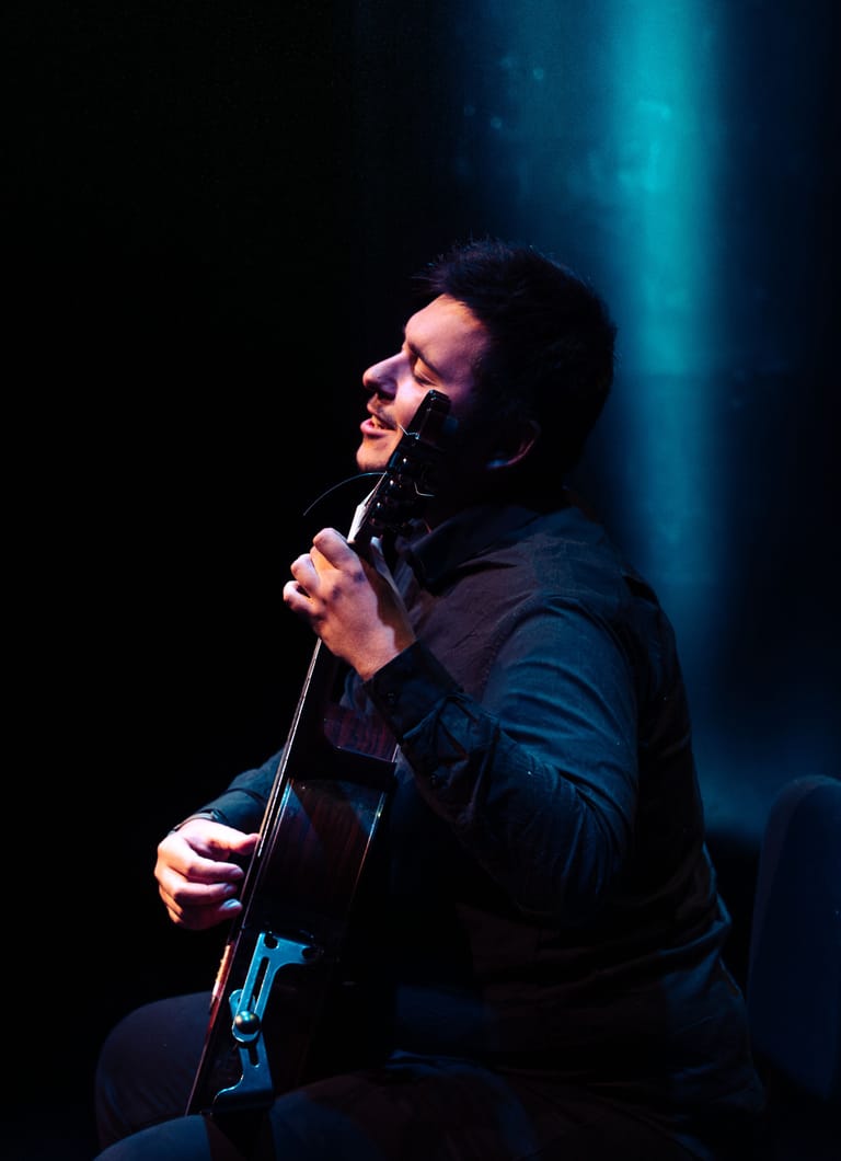 Performer singing into a microphone while holding an electric guitar, lit by blue stage spotlights.