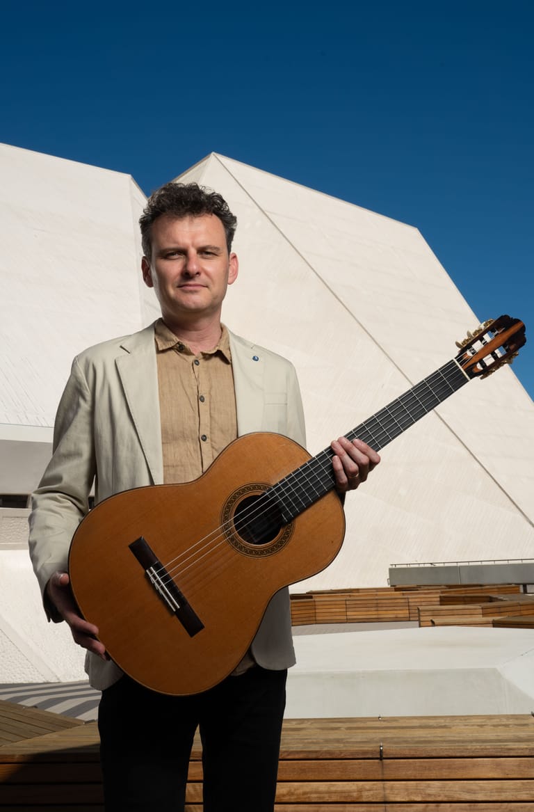 Musician holding a classical guitar against a modern white building and clear blue sky.