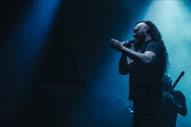 Musician with long dark hair singing into microphone on stage bathed in blue spotlight lighting.