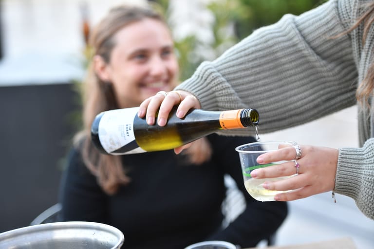 Person in grey knit pouring white wine into a glass at an outdoor gathering.