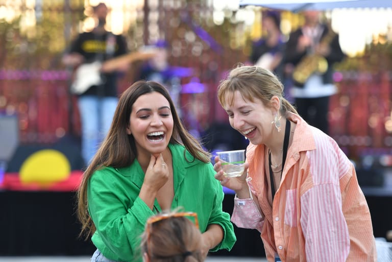 Two women laugh together at an outdoor event with festival atmosphere and crowd in background.