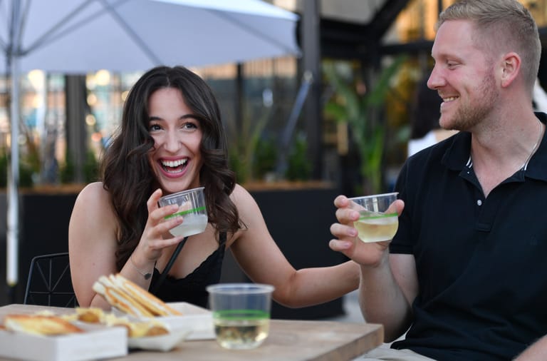Two people toast with glasses of clear beverage at an outdoor dining table with food and plants in the background.