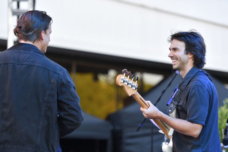 Two musicians in blue shirts conversing outdoors, one holding an acoustic guitar.