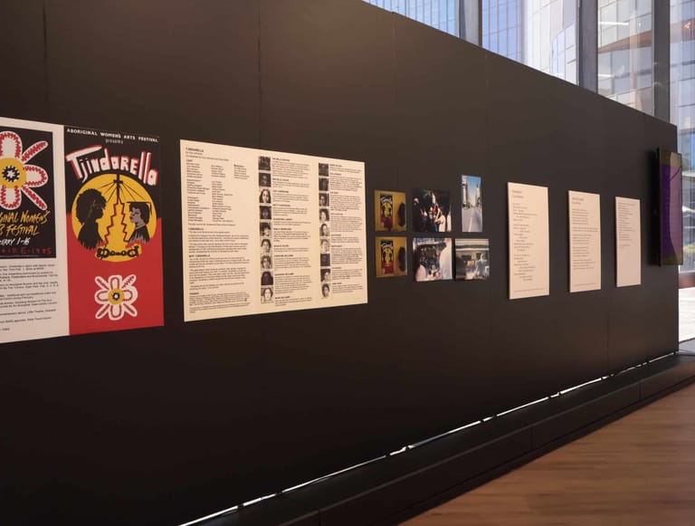 Exhibition wall displaying vintage posters, archival documents and black-and-white photographs on dark panelling.