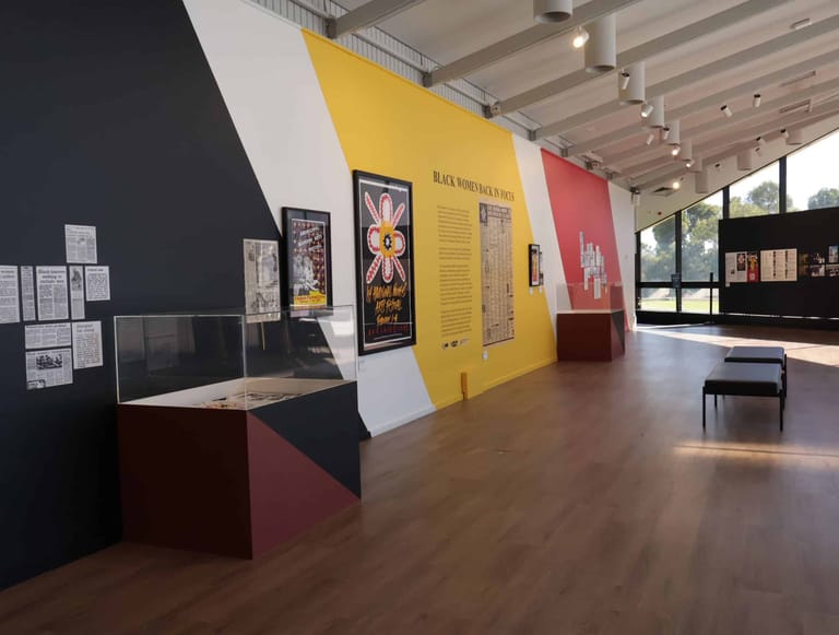 Modern gallery interior with yellow and red accent walls displaying artwork and exhibition materials on dark and light surfaces.