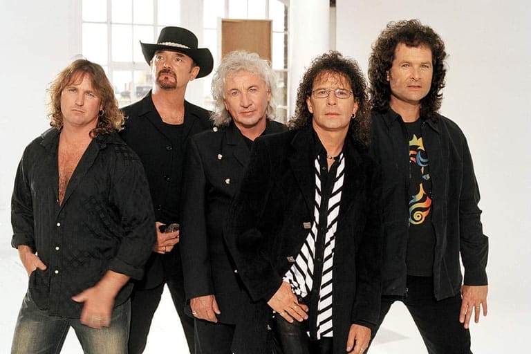 Five musicians in black jackets stand together against a white brick wall backdrop.