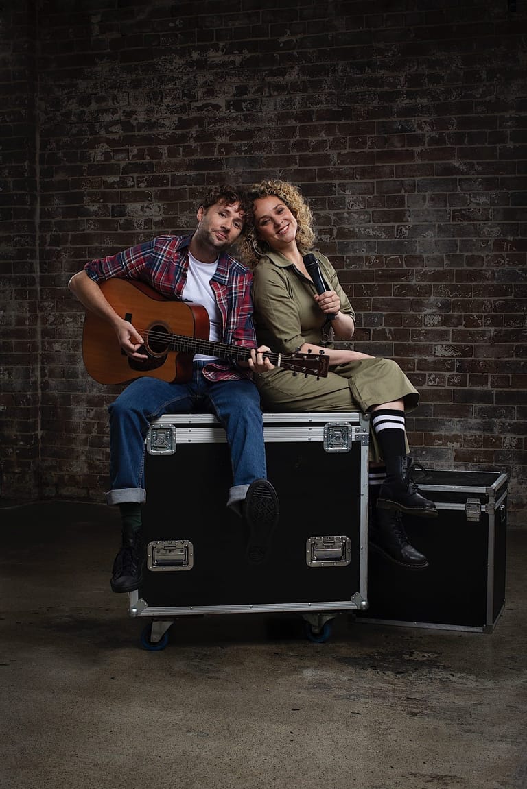 Two musicians sit on black flight cases against a brick wall, one holding an acoustic guitar and the other a keyboard instrument.