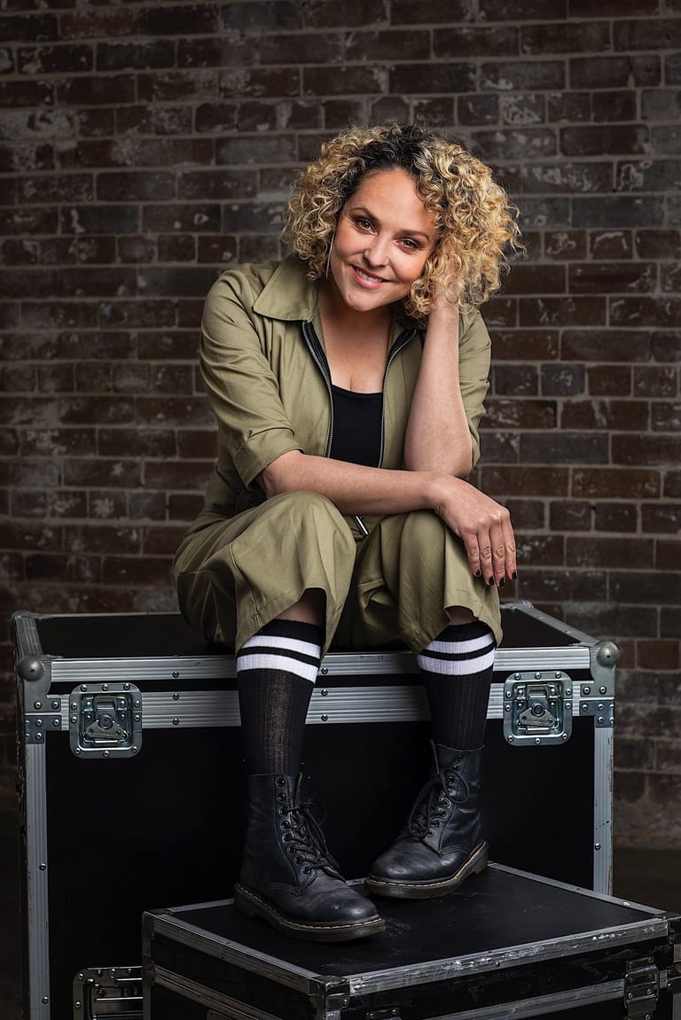 Person with curly blonde hair seated on a flight case against a brick wall, wearing olive jacket and black boots.