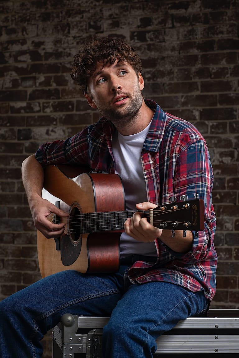 Musician holding an acoustic guitar, wearing a red plaid shirt against an exposed brick wall.