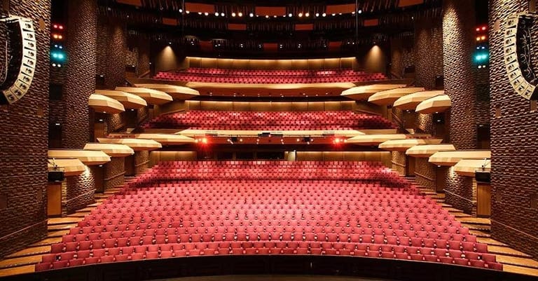 Empty theatre auditorium with red seats, gold railings, and warm stage lighting creating an inviting venue interior.