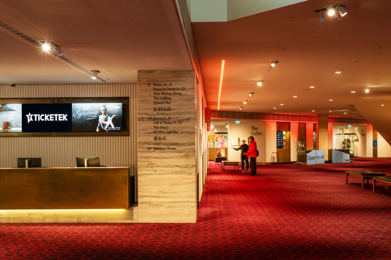 Cinema foyer with ticketing desk, red carpet, and illuminated entrance doors under warm overhead lighting.