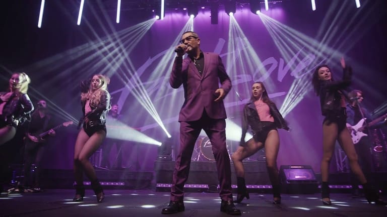 Performer in burgundy suit centre stage surrounded by dancers, bathed in purple spotlights and geometric light beams.