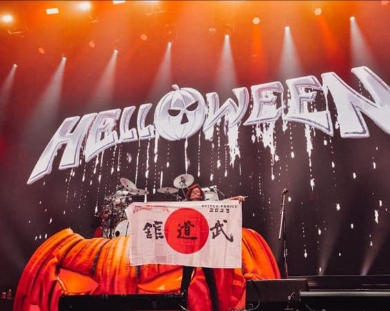 Helloween logo projected above a drummer beneath stage spotlights during a concert performance.