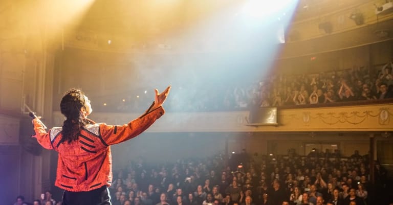 Performer in a red jacket with arms outstretched addressing an audience in a theatre lit by stage spotlights.