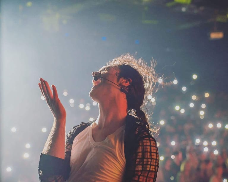 Performer with blonde hair in plaid shirt singing with hand raised, bathed in warm stage lighting and bokeh lights.