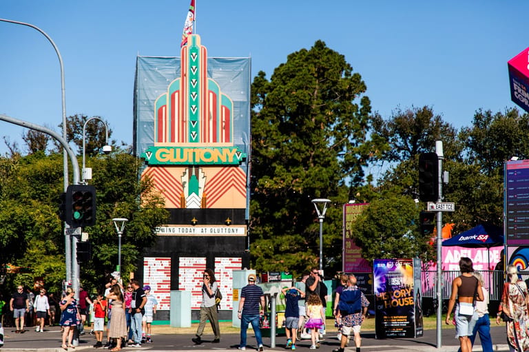Crowds gather beneath a tall art deco cinema marquee on a sunny day surrounded by green trees.