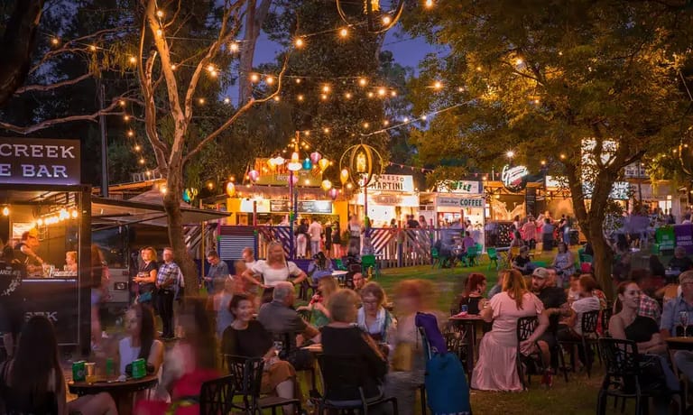 Twilight scene of an outdoor festival with crowds gathered under string lights and trees, food vendors visible at left.