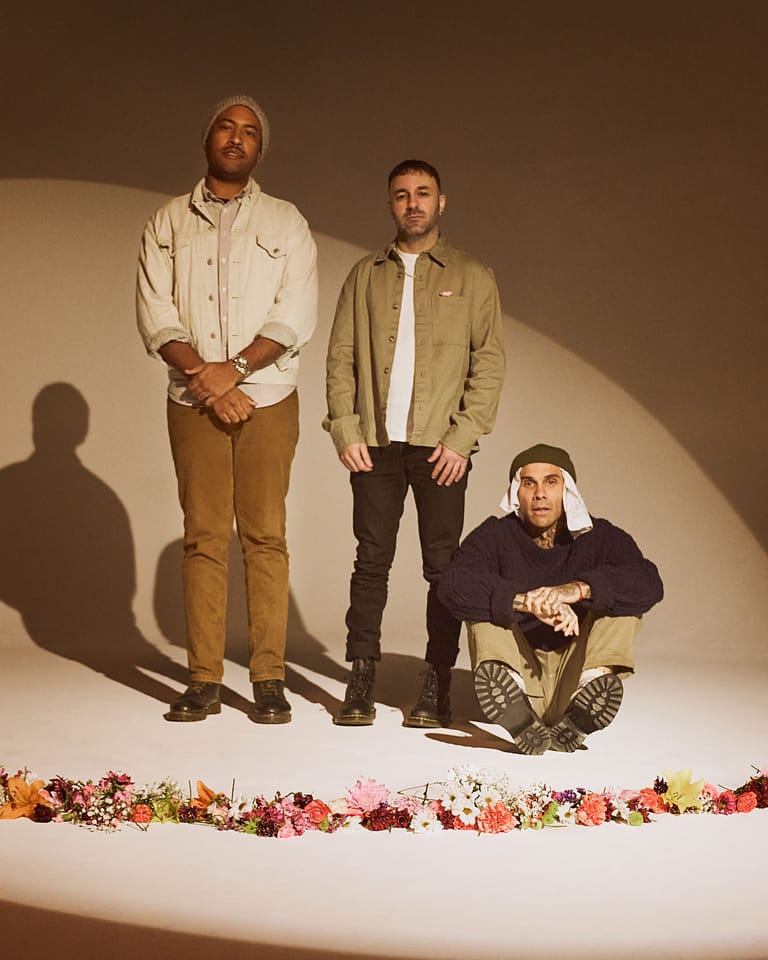Three male performers posed against a cream backdrop with floral garland, one seated and two standing in casual attire.