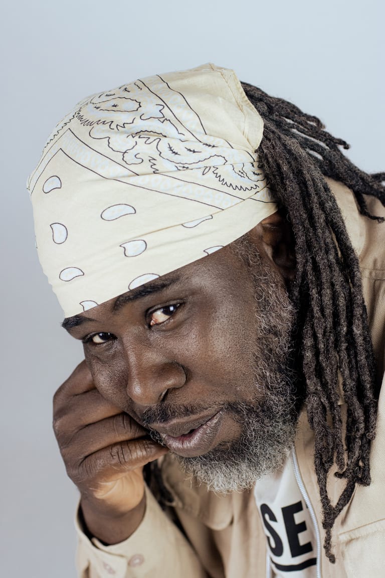 Man with dreadlocks and patterned headwrap rests chin on hand against neutral background.