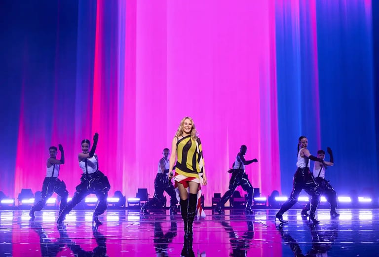Performer in yellow jacket centre stage with dancers flanking either side beneath vivid magenta and blue neon lighting.