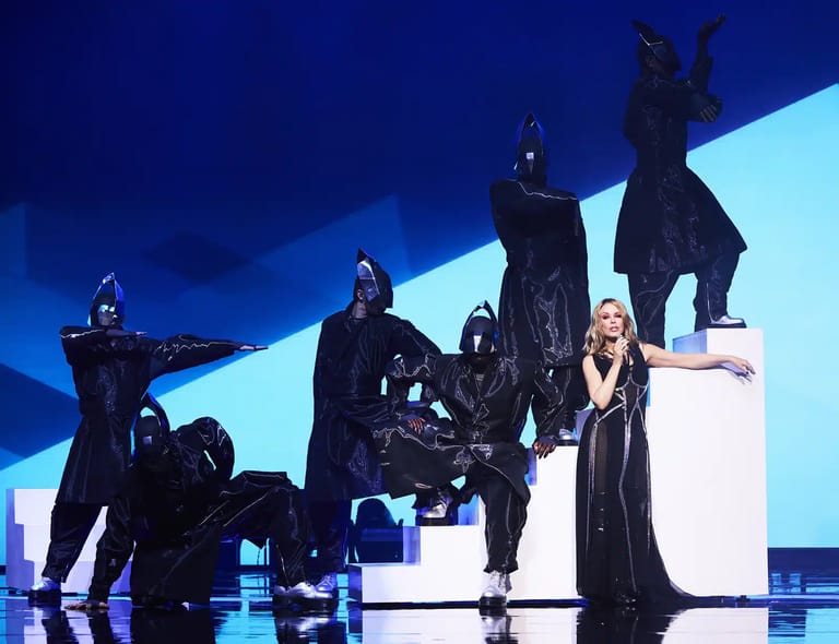Performers in dark theatrical costume posed on an illuminated stage with dramatic blue lighting and sculptural set pieces.