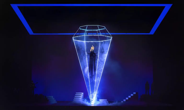 Performer standing illuminated inside a glowing blue geometric structure on a dark stage.