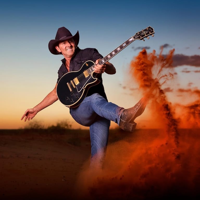 Guitarist in cowboy hat and jeans kicks up red dust at sunset, holding black and gold electric guitar.