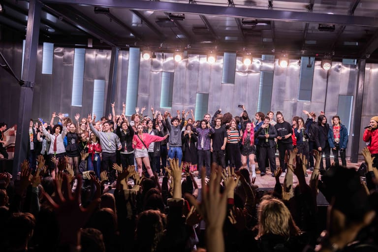 Large crowd of audience members with raised hands and phones facing a brightly lit stage with performers and vertical lighting installations.