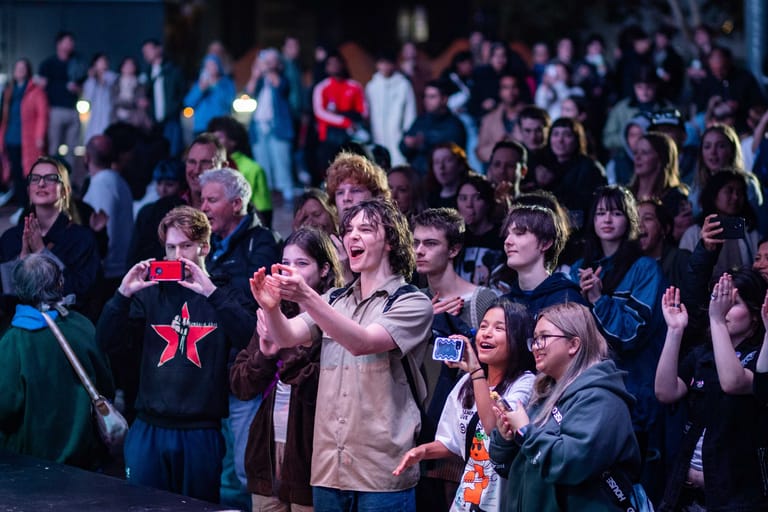 Crowd of audience members clapping and cheering at a live entertainment event, bathed in stage lighting.