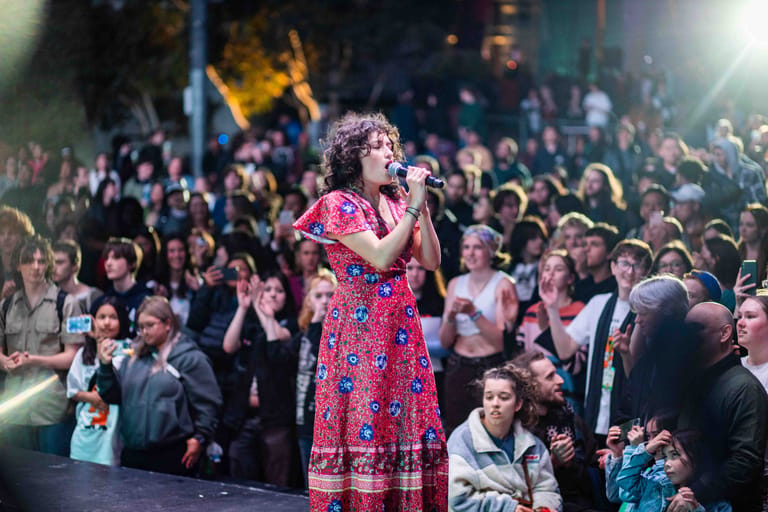 Performer in a vibrant pink patterned dress singing to a crowded venue audience lit by stage lighting.