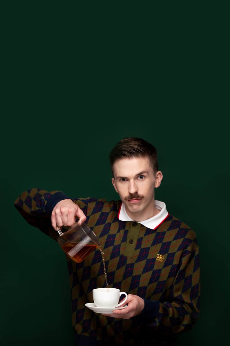 Man in patterned sweater pouring tea into white cup against deep green background.