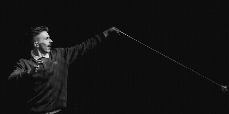Man in black jacket holding microphone on stage, arm extended dramatically against dark background.